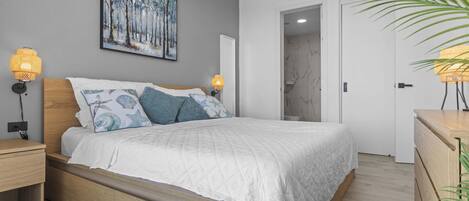 Une chambre à coucher comprenant un lit, des lampes de chevet, une table de nuit, une tête de lit en bois, un tableau accroché au mur et une porte donnant sur une autre pièce.