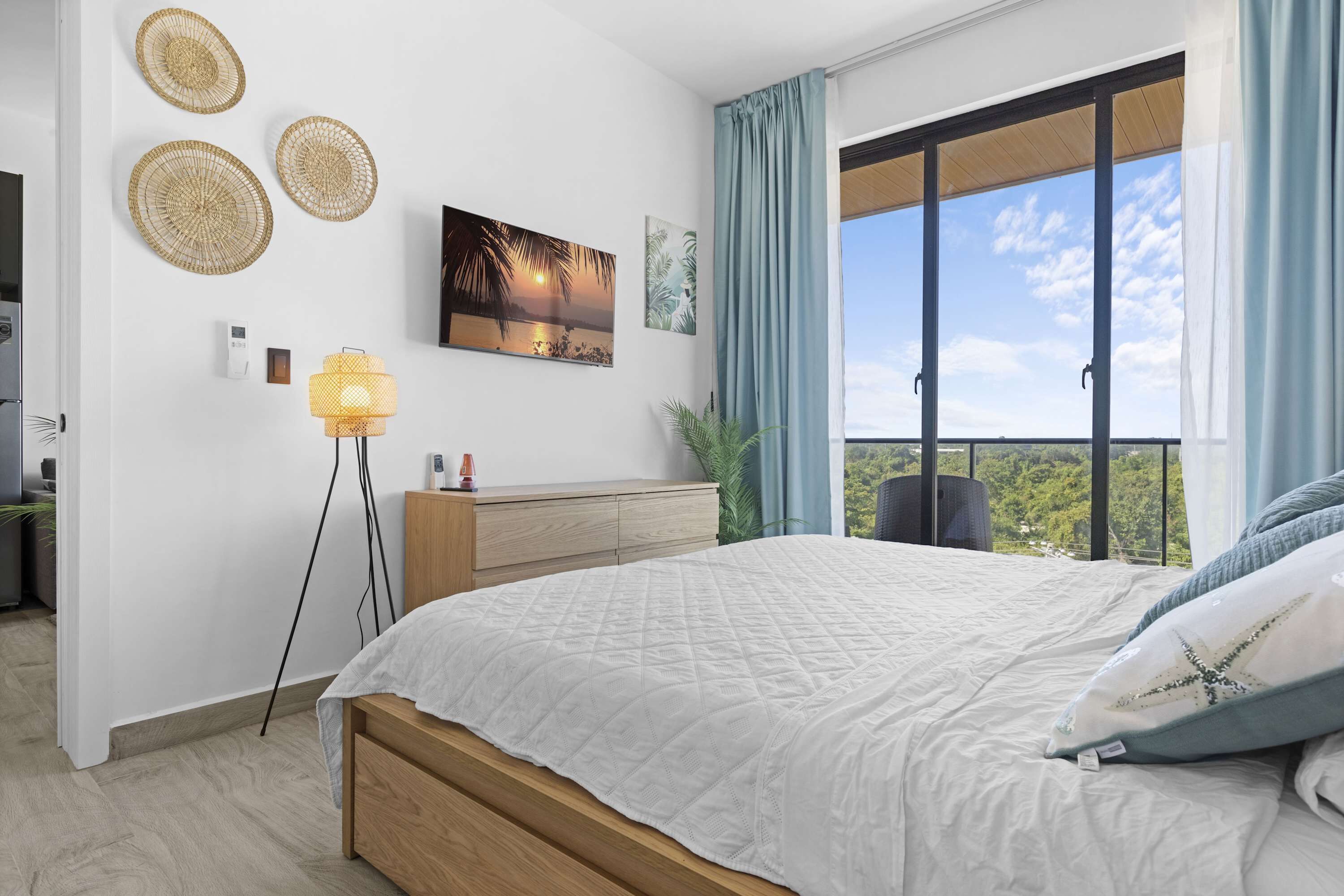 A bedroom with a bed, a bedside table, a lamp, a wall-mounted artwork, and a view of the outdoors.