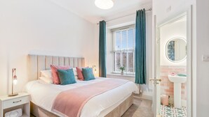 Une chambre à coucher comprenant un lit, une table de chevet, une lampe, une fenêtre avec des rideaux et une vue sur la salle de bain.