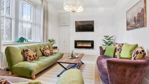 Un salon avec un canapé vert, un fauteuil rouge, une cheminée et une grande fenêtre équipée de stores.