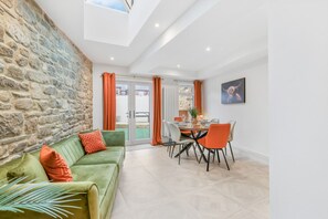 A modern living room with a stone wall, a green sofa with orange cushions, a dining area with orange chairs, and a view through a glass door. - Church View - North East Escapes (Lanchester)