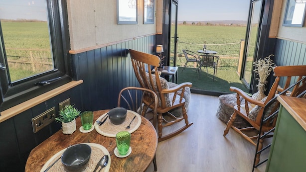 Een eethoek met een tafel voor twee personen, houten stoelen en uitzicht op een groen veld door openslaande deuren.