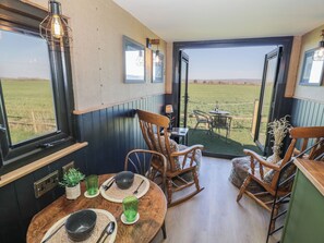 A dining area with a table set for two, wooden chairs, and a view of a green field through open doors. - Gigi's Horsebox (Evesham)