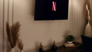 Un salon moderne avec un téléviseur à écran plat fixé au mur, deux vases contenant des plantes séchées, une petite statue et une plante en pot posée sur une console en bois.