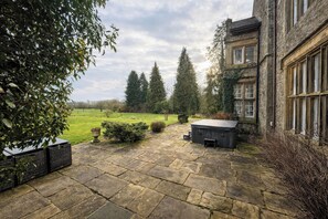 A stone patio with a hot tub and seating area, surrounded by trees and a view of a green field.