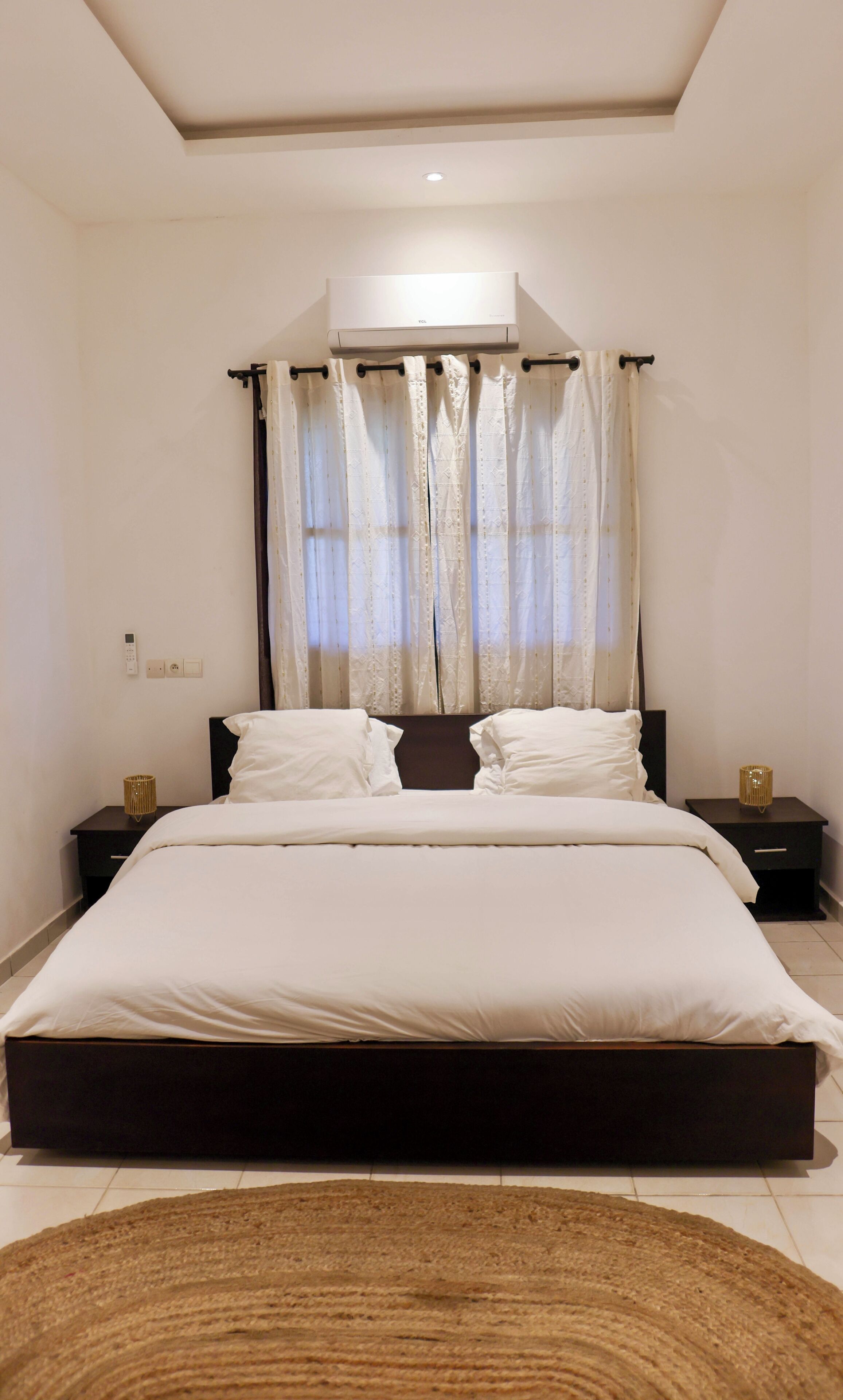 A bedroom with a large bed, a ceiling-mounted air conditioner, a bedside lamp, and a nightstand.