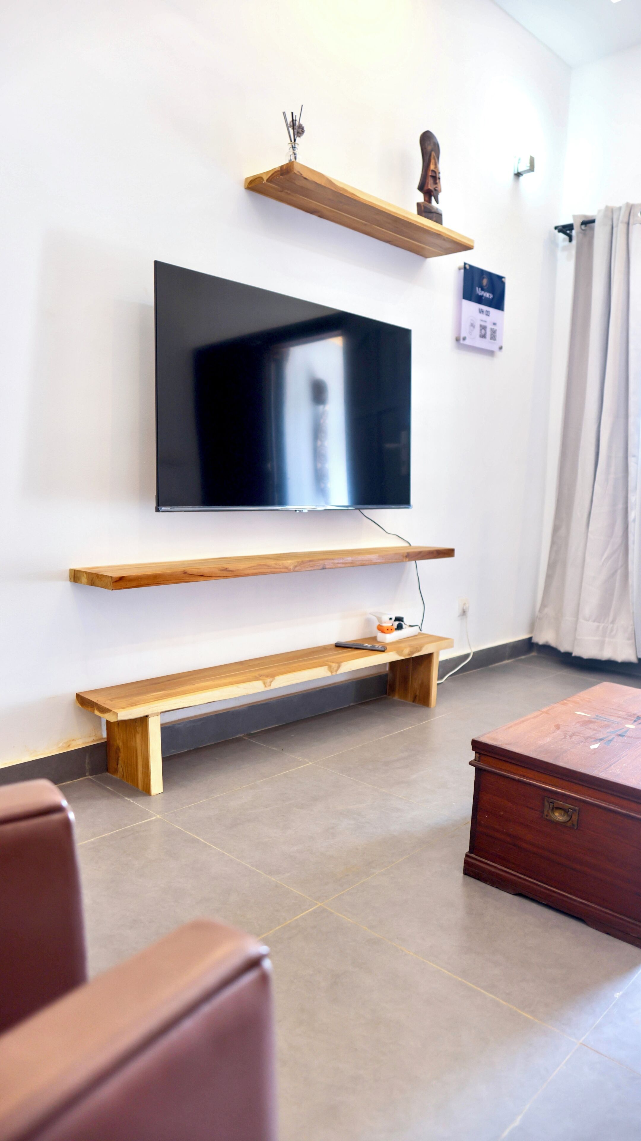 Una sala de estar con un televisor de pantalla plana montado en la pared, estantes de madera, un banco, una mesita y un armario.