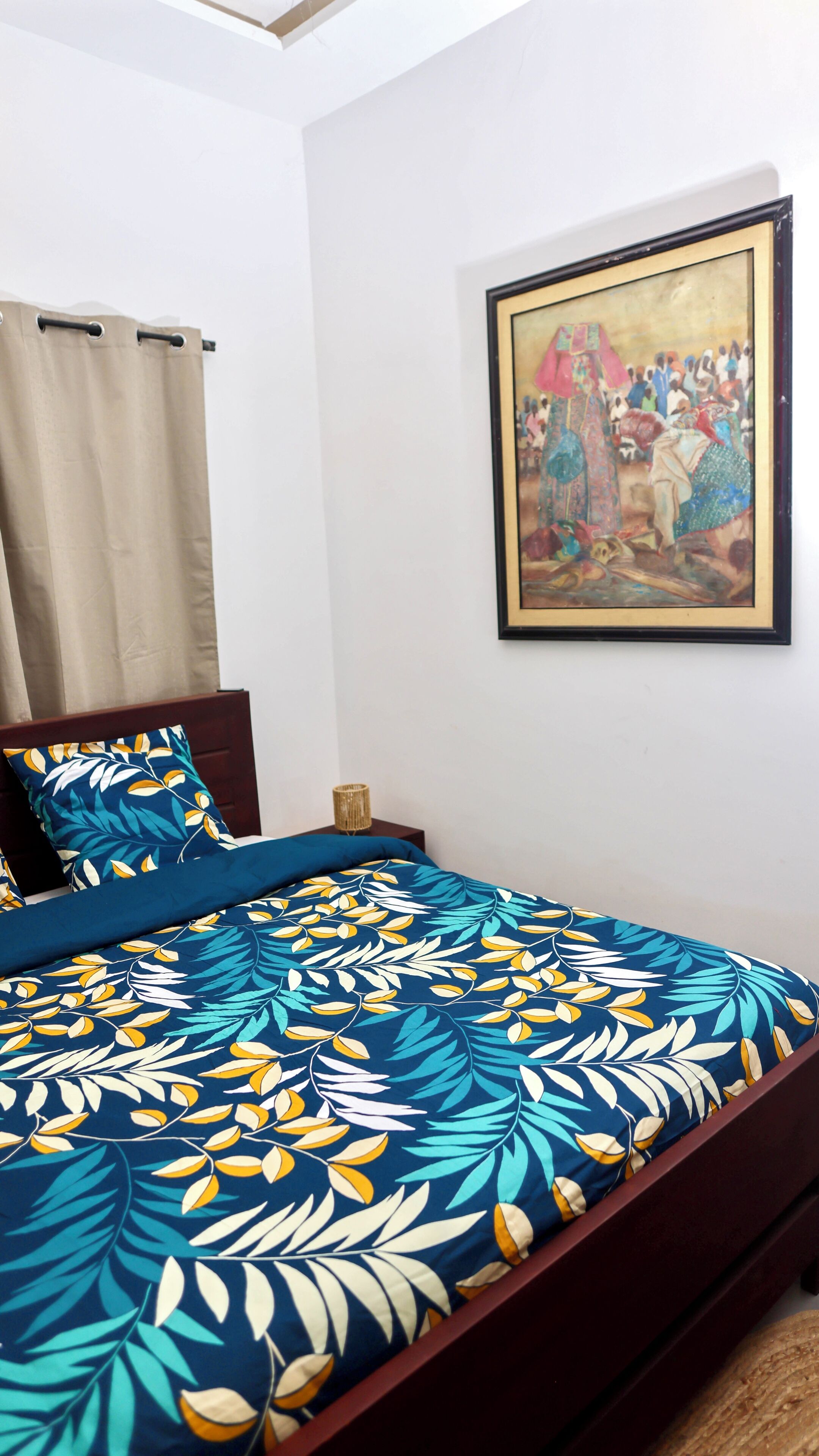 Een slaapkamer met een bed, een nachtkastje, een lijst met een schilderij en een raam met gordijnen.