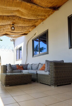 A covered outdoor seating area with a wicker sofa, a ceiling fan, and a window with a view of palm trees. - MAYORA Villas & Suites (Lomé)