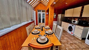 Un comedor con mesa de madera dispuesta para seis personas, rodeada de sillas blancas. A la derecha se ve una cocina con lavadora y armarios.