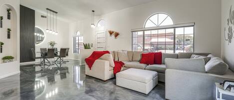 A spacious living room with a large sectional sofa, a dining area with a glass table, and a modern kitchen visible through an open door.