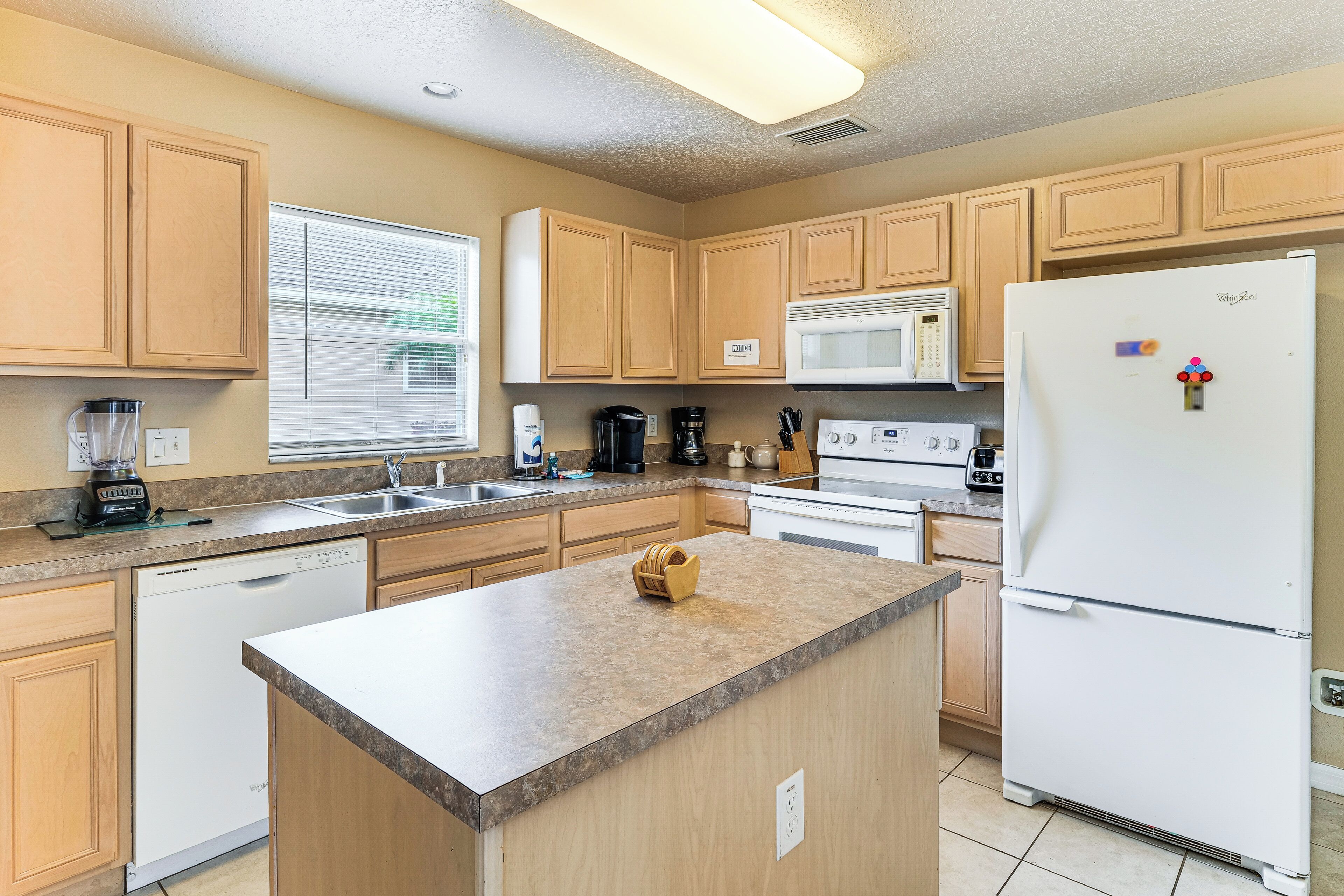 A kitchen with wooden cabinets, a built-in microwave, a refrigerator, a dishwasher, and a window with blinds.
