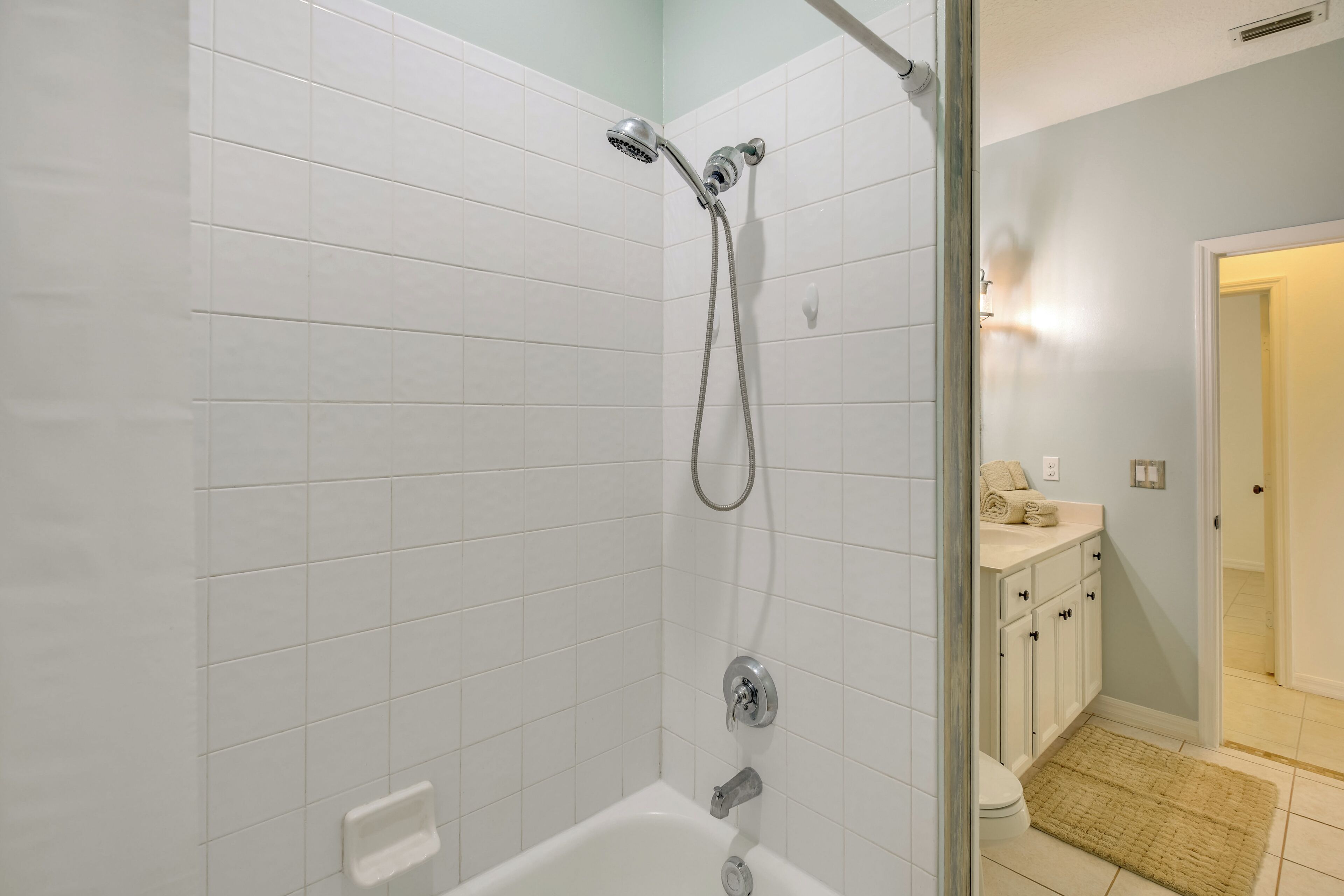 Ein Badezimmer mit weißen Fliesen, einer Dusche mit Handbrause, einer Badewanne und einem kleinen Waschbecken mit einem Handtuch.