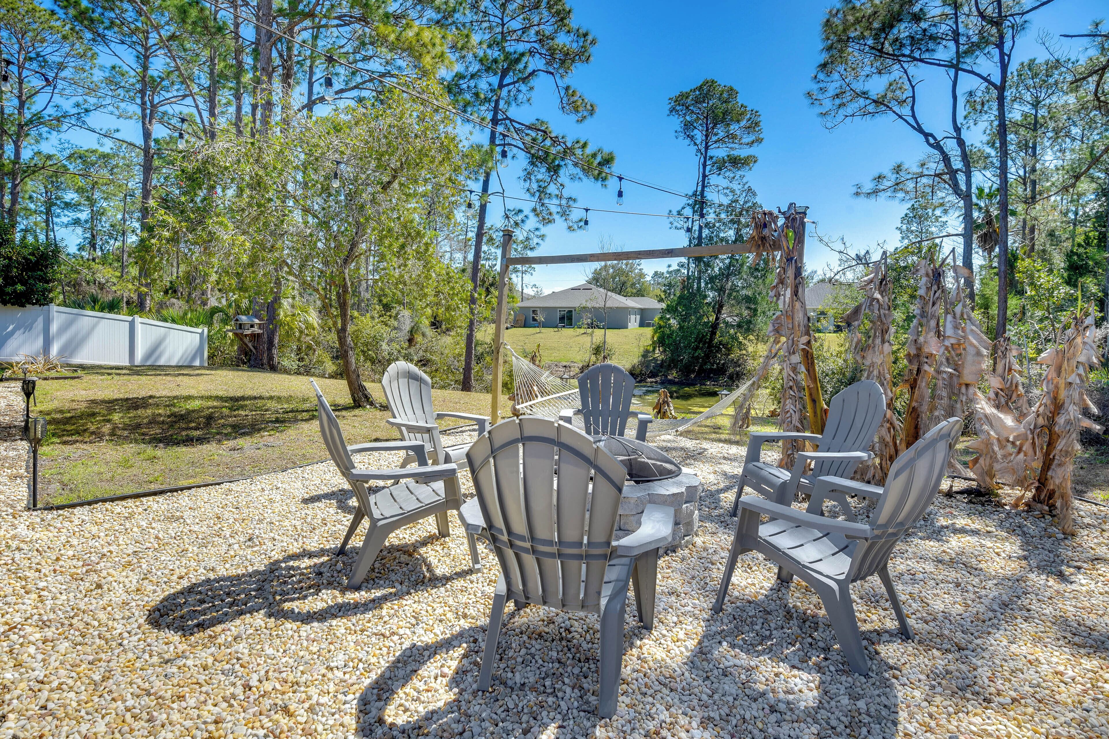 An outdoor seating area with Adirondack chairs and a stone fire table, surrounded by trees and a house in the background.