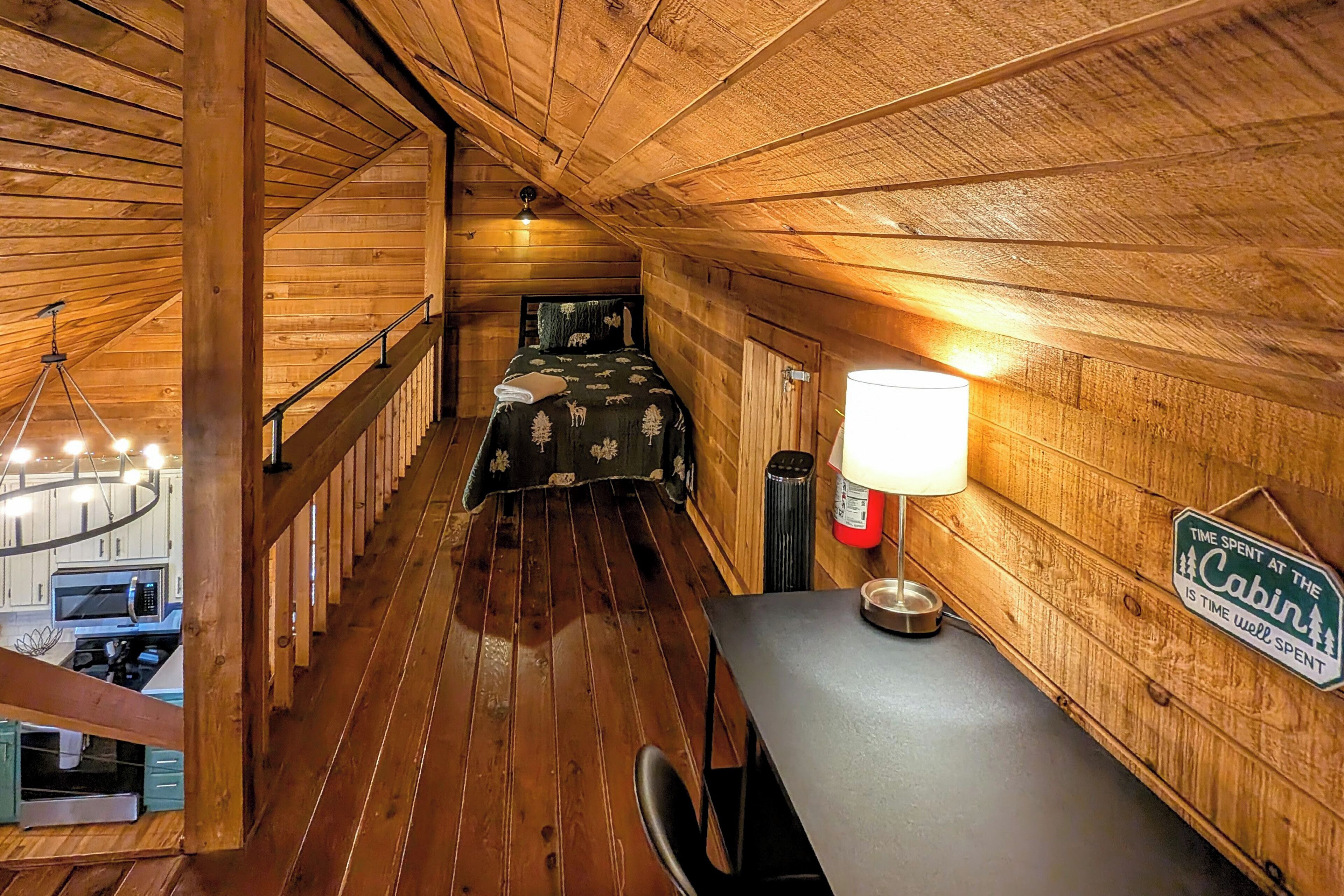 A cabin interior with a wooden floor, a bed with a patterned bedspread, a desk with a lamp, a sign on the wall, and a staircase leading to another room.