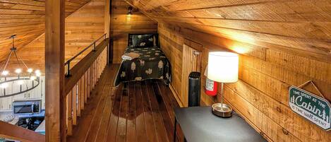 A cabin interior with a wooden floor, a bed with a patterned bedspread, a desk with a lamp, a sign on the wall, and a staircase leading to another room.
