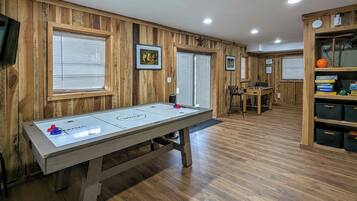 A room with wooden paneled walls, a shuffleboard table, a billiards table, and a bookshelf with storage boxes.