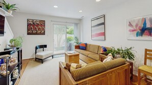 A modern living room with a sofa, armchair, and a wooden bench. There are two potted plants, a bookshelf, and a large window with a view of greenery outside. - 5 Blocks to Shore! Rehoboth Beach Family Escape (Rehoboth Beach)