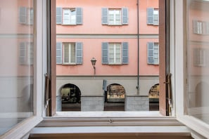 Une vue par la fenêtre sur un immeuble doté de nombreuses fenêtres et de volets, un lampadaire et une arche en pierre.