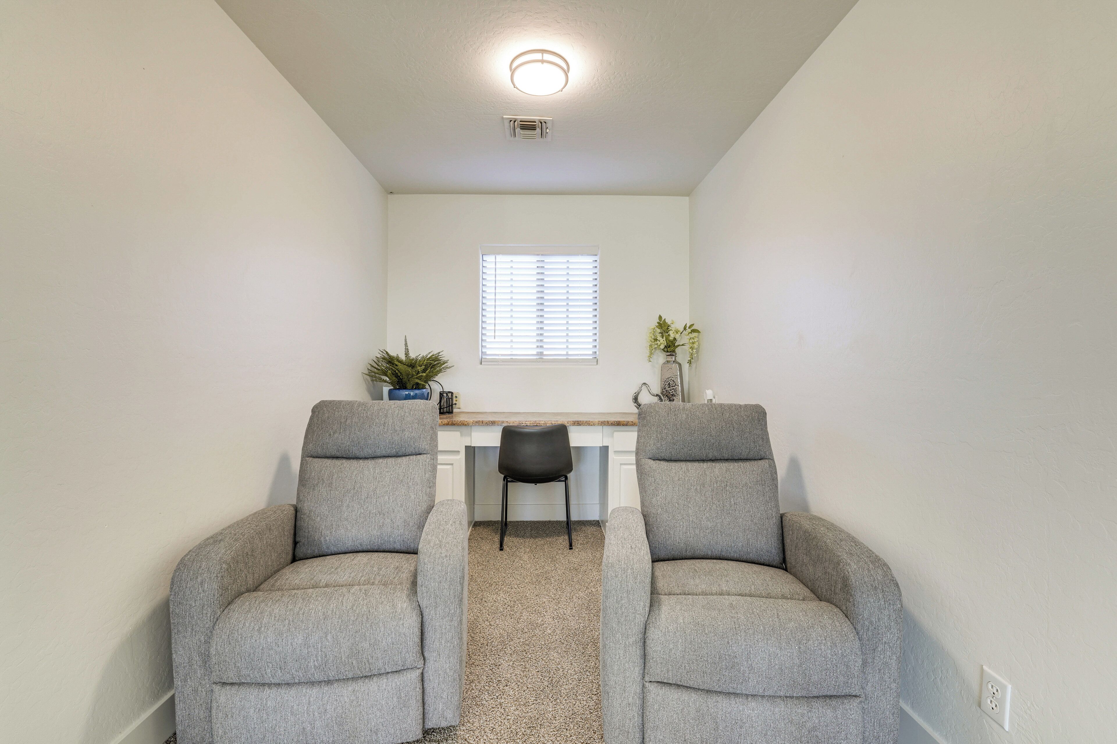 A room with two grey armchairs facing a desk with a chair, a window with blinds, and a ceiling light.