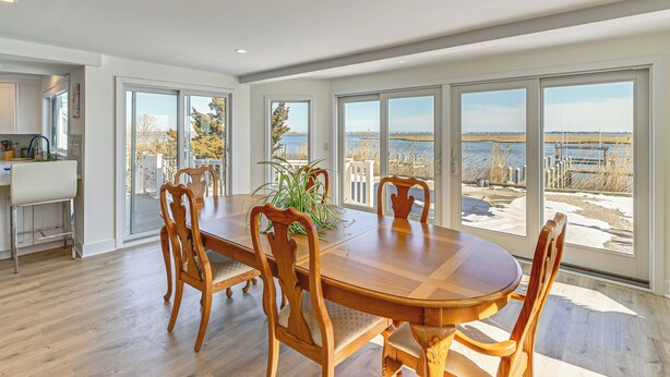A dining area with a wooden table and chairs, large windows with a view of the water and trees, and a kitchen area with a sink and countertop. - Private Pool, Dock & Beach: Stafford Township Gem (Manahawkin)