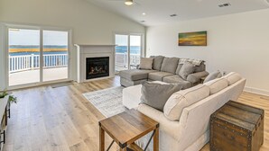 A spacious living room with a large sectional sofa, a fireplace, and a view of the ocean through large sliding doors. - Private Pool, Dock & Beach: Stafford Township Gem (Manahawkin)