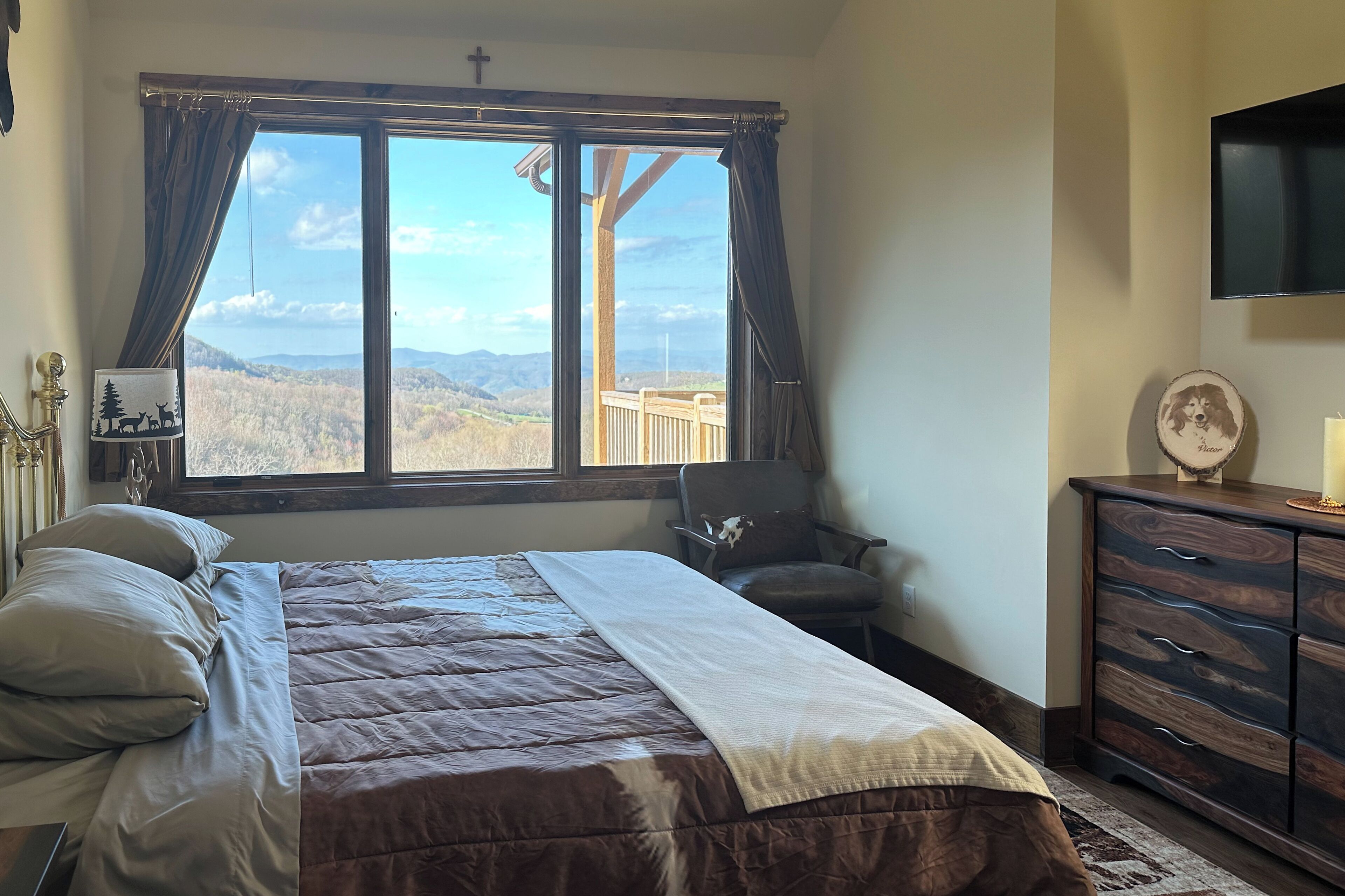 A bedroom with a large window, a bed with a brown quilt, a wooden dresser, and a small chair.