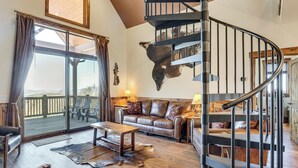 Un salon spacieux avec un canapé en cuir, une table basse en bois et une grande fenêtre avec des rideaux. On aperçoit un escalier en colimaçon au fond.