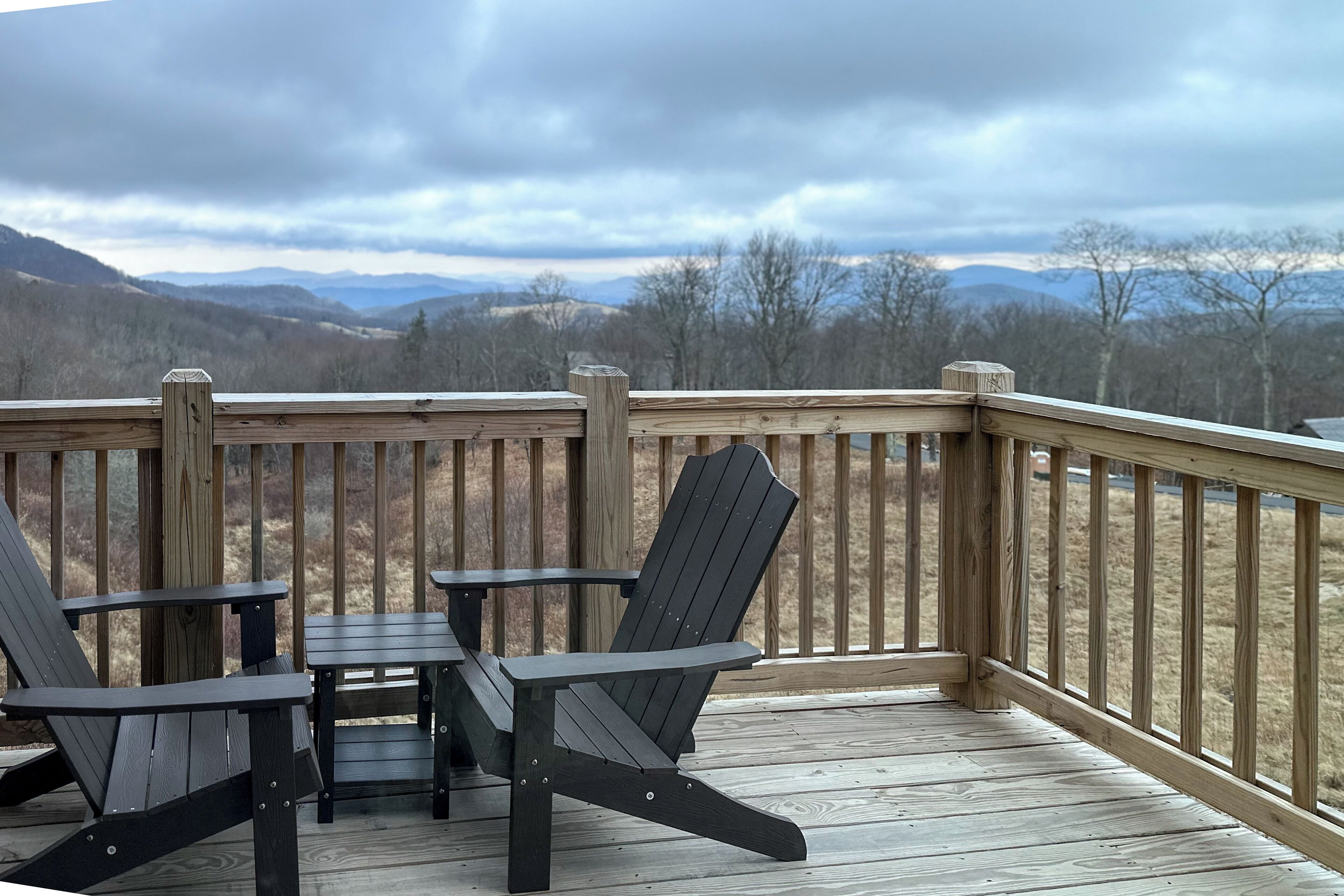 A wooden deck with two Adirondack chairs and a table, overlooking a mountainous landscape.