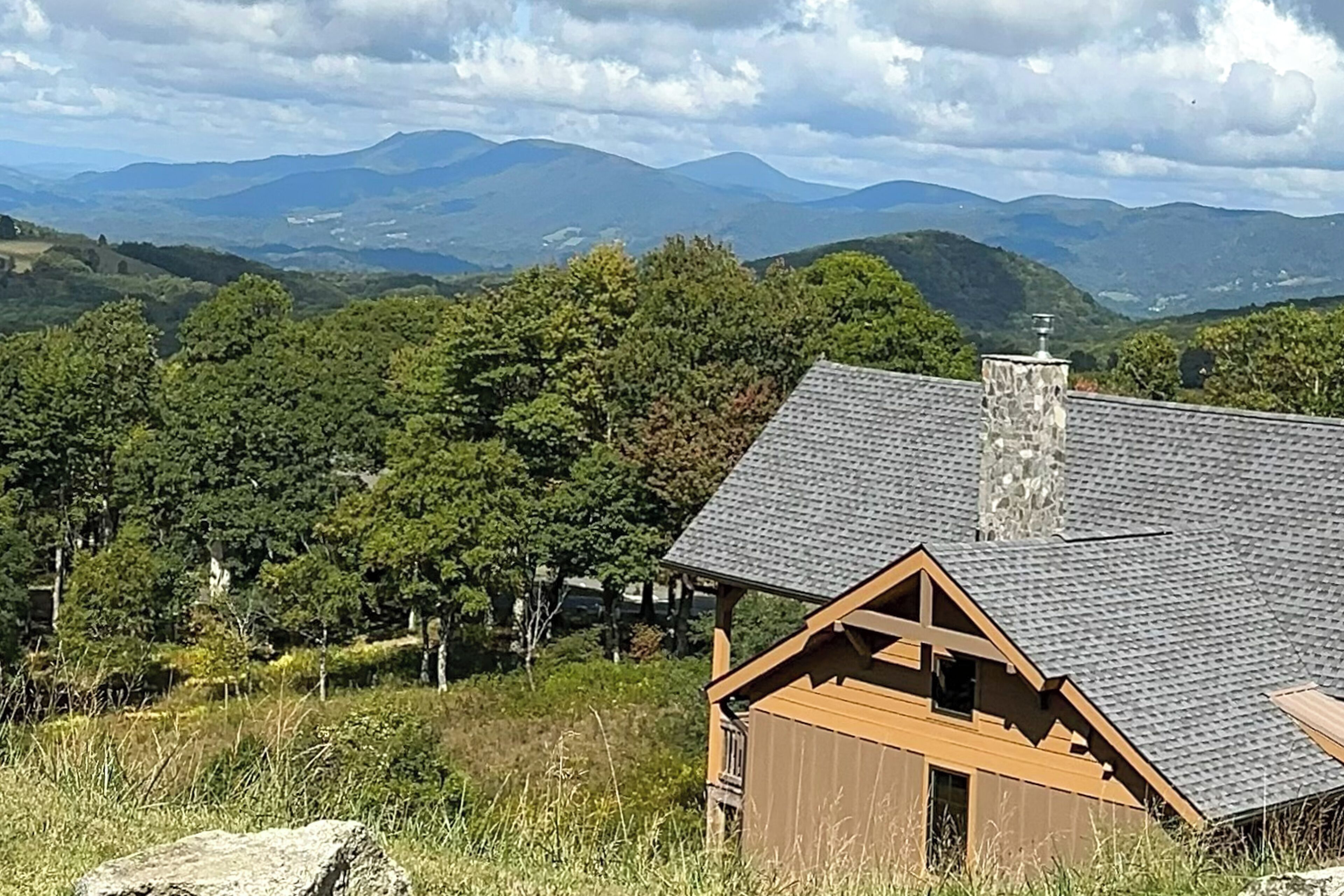 A cabin with a sloped roof, surrounded by lush greenery and mountains in the background.