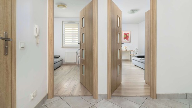 A hallway with wooden doors leading to a bedroom with a bed, a bedside table, and a window with blinds. - Functional Apartment with Balcony | Quiet Neighborhood (Kraków)