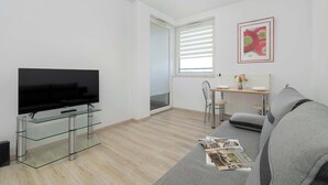 A modern living room with a flat-screen TV on a glass stand, a grey sofa, a dining area with a table and chairs, and a window with blinds. - Functional Apartment with Balcony | Quiet Neighborhood (Kraków)