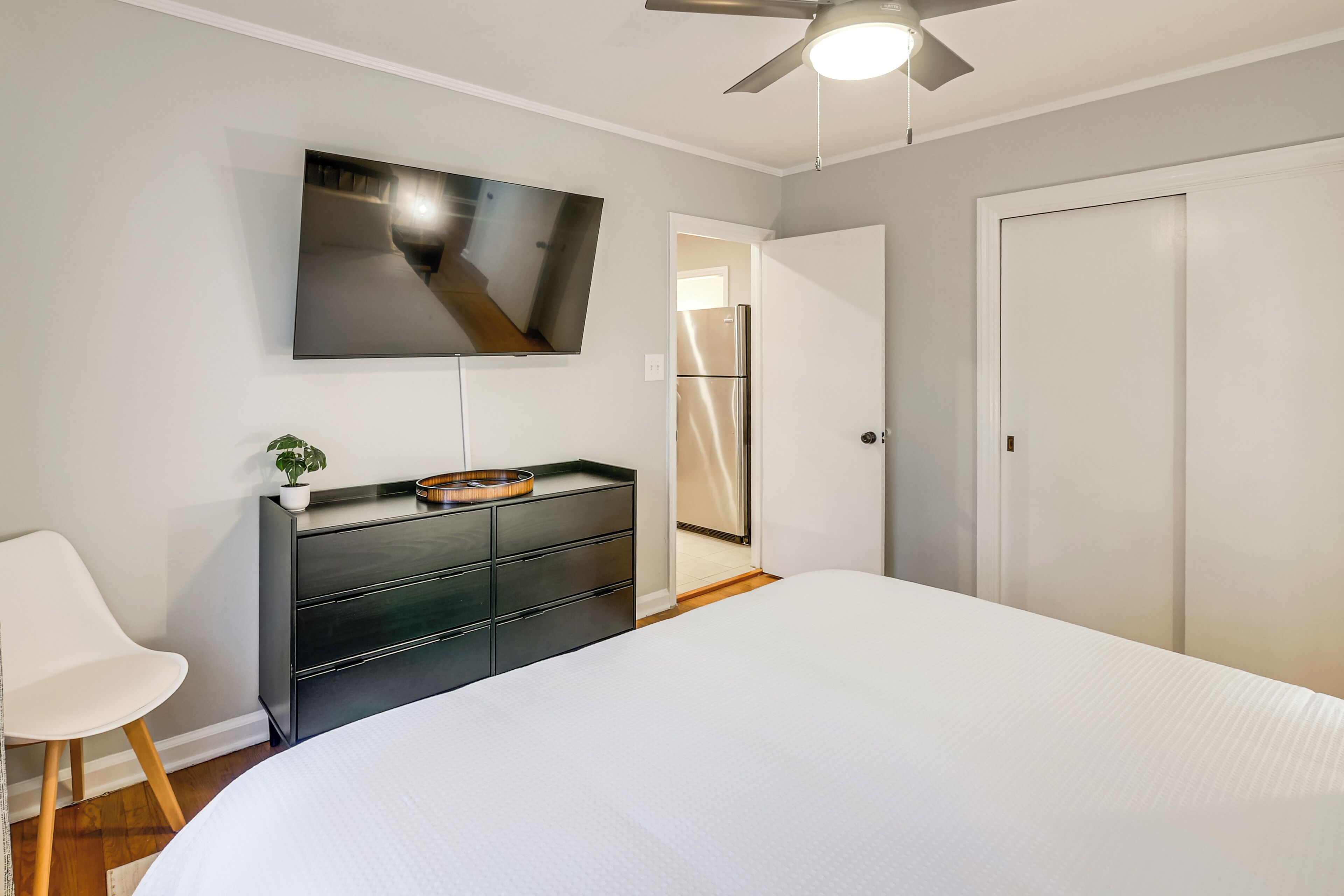 A bedroom with a bed, a TV mounted on the wall, a dresser, a chair, and a door leading to another room.
