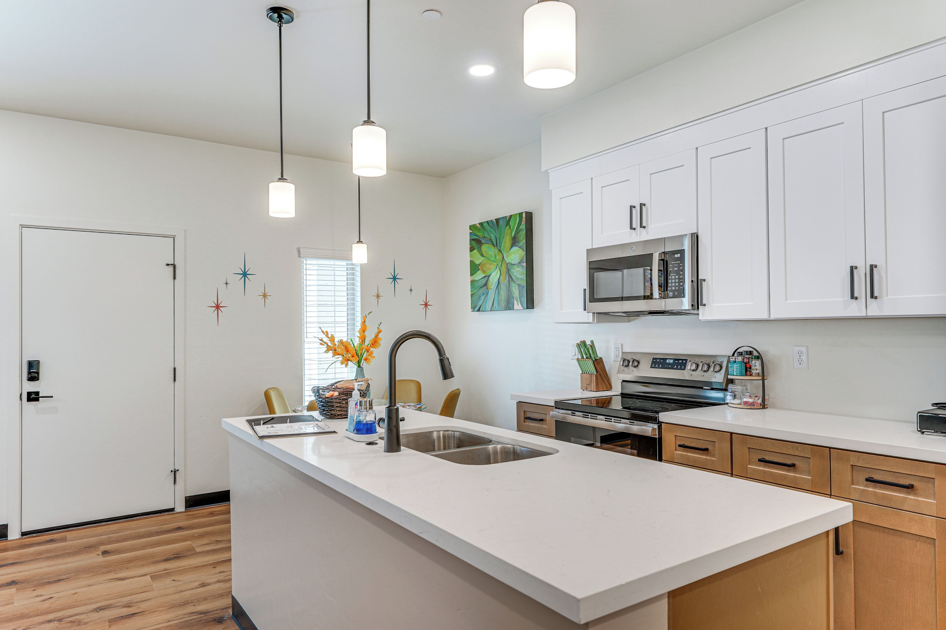 A modern kitchen with white cabinetry, a central island with a sink, stainless steel appliances, pendant lighting, and a decorative wall piece.