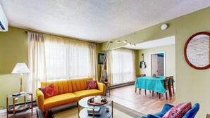 Uma sala de estar com um sofá amarelo, poltronas azuis, uma mesa de centro de vidro e um espaço de refeições com toalha de mesa turquesa.