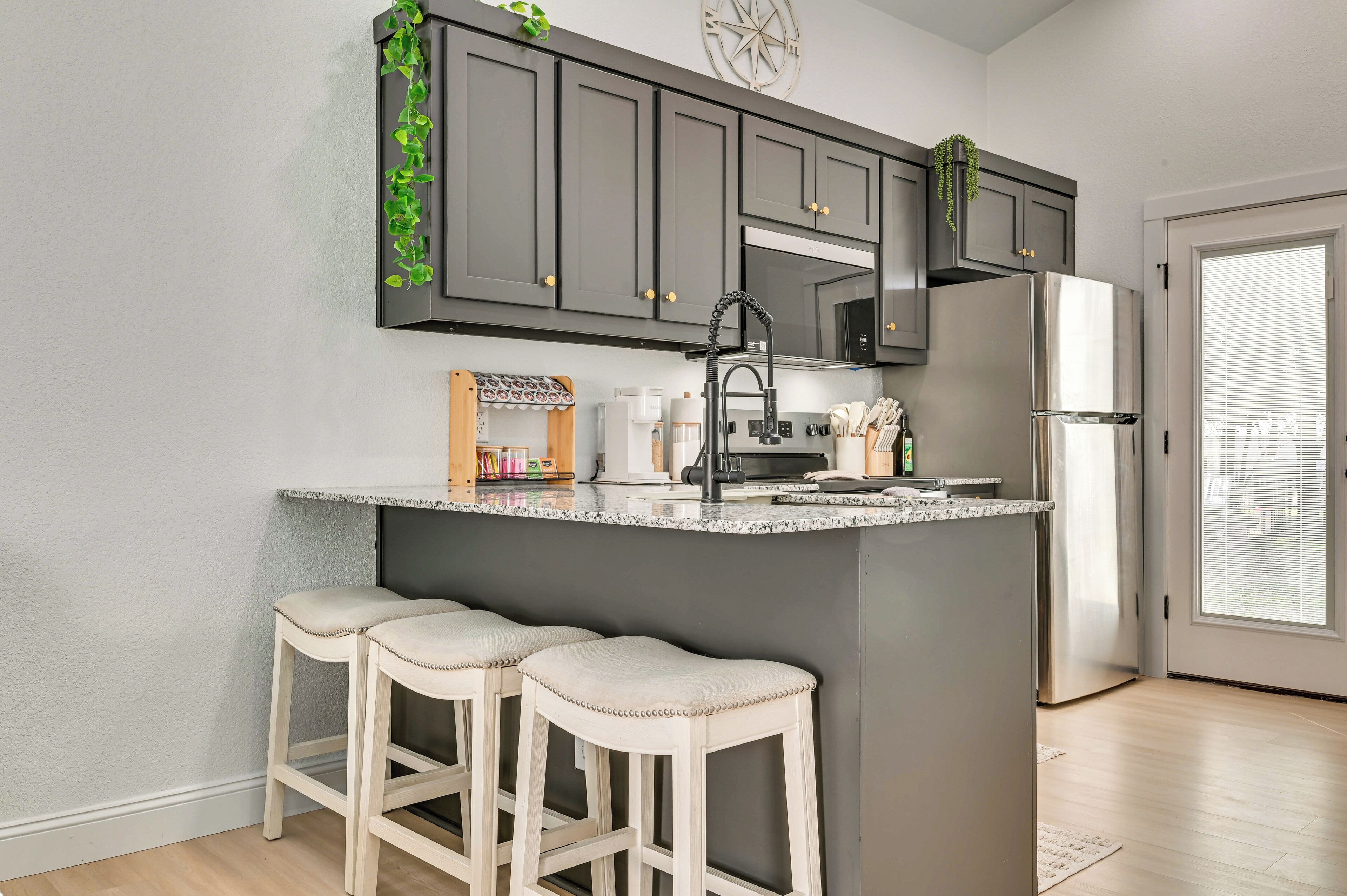 A kitchen with dark grey cabinets, a stainless steel refrigerator, a granite countertop, and two white bar stools.