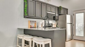 Una cocina con armarios gris oscuro, un refrigerador de acero inoxidable, una encimera de granito y dos taburetes blancos para bar.
