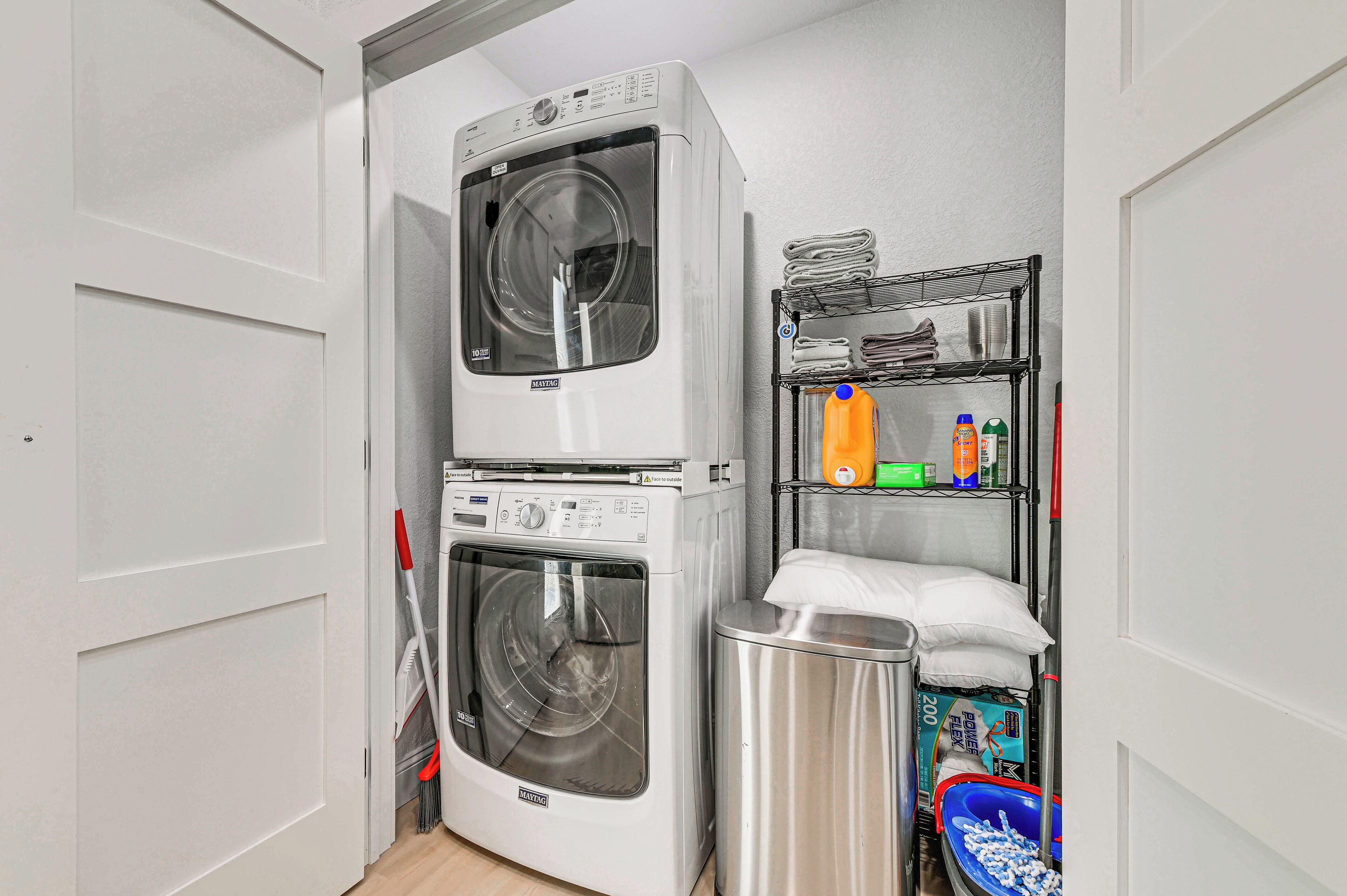 A laundry room with a stacked washer and dryer, a metal shelf with folded towels, cleaning supplies, and a trash bin.