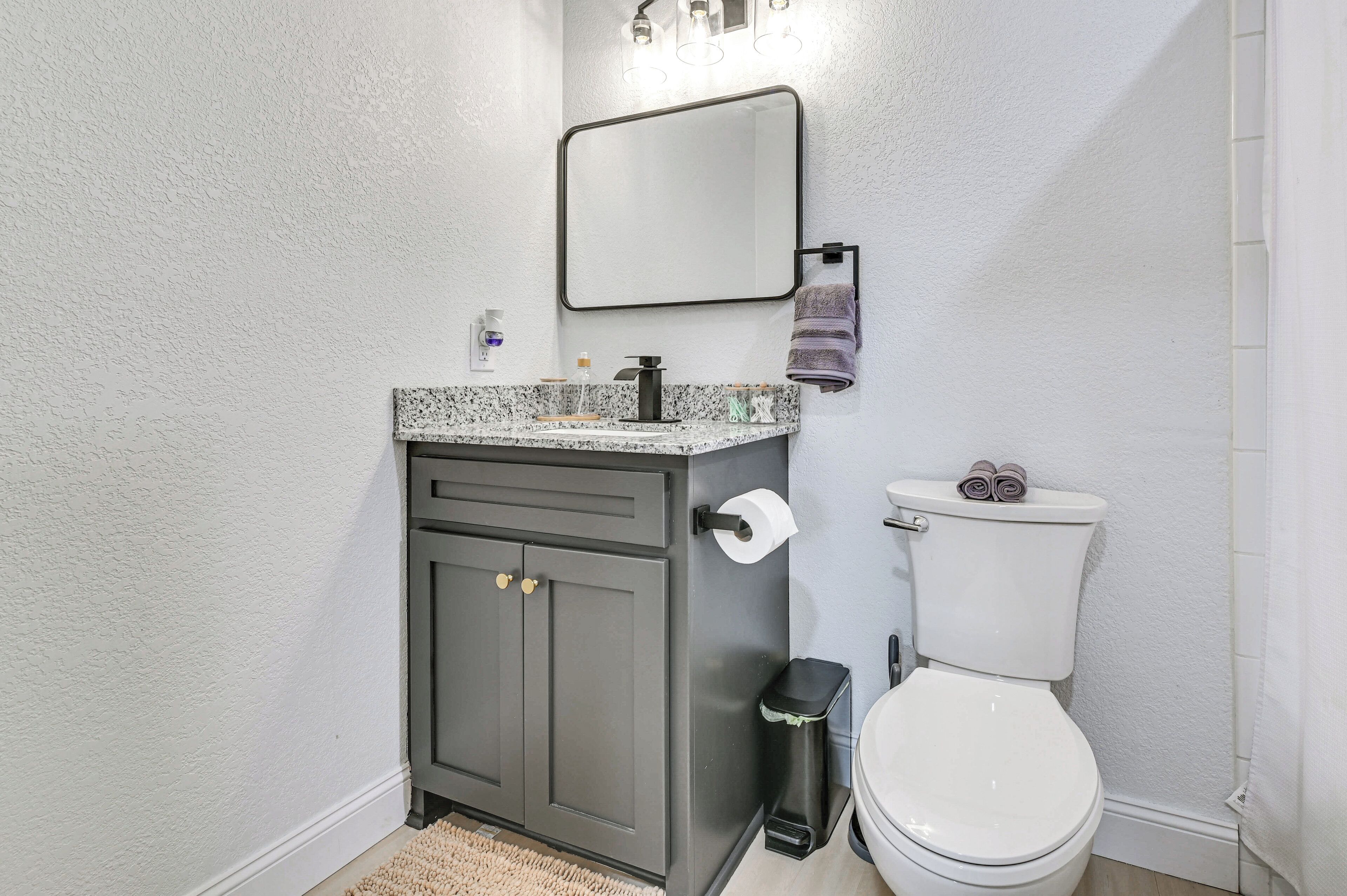 A bathroom with a white toilet, a dark grey cabinet with gold handles, a marble countertop, a mirror, and a towel rack.