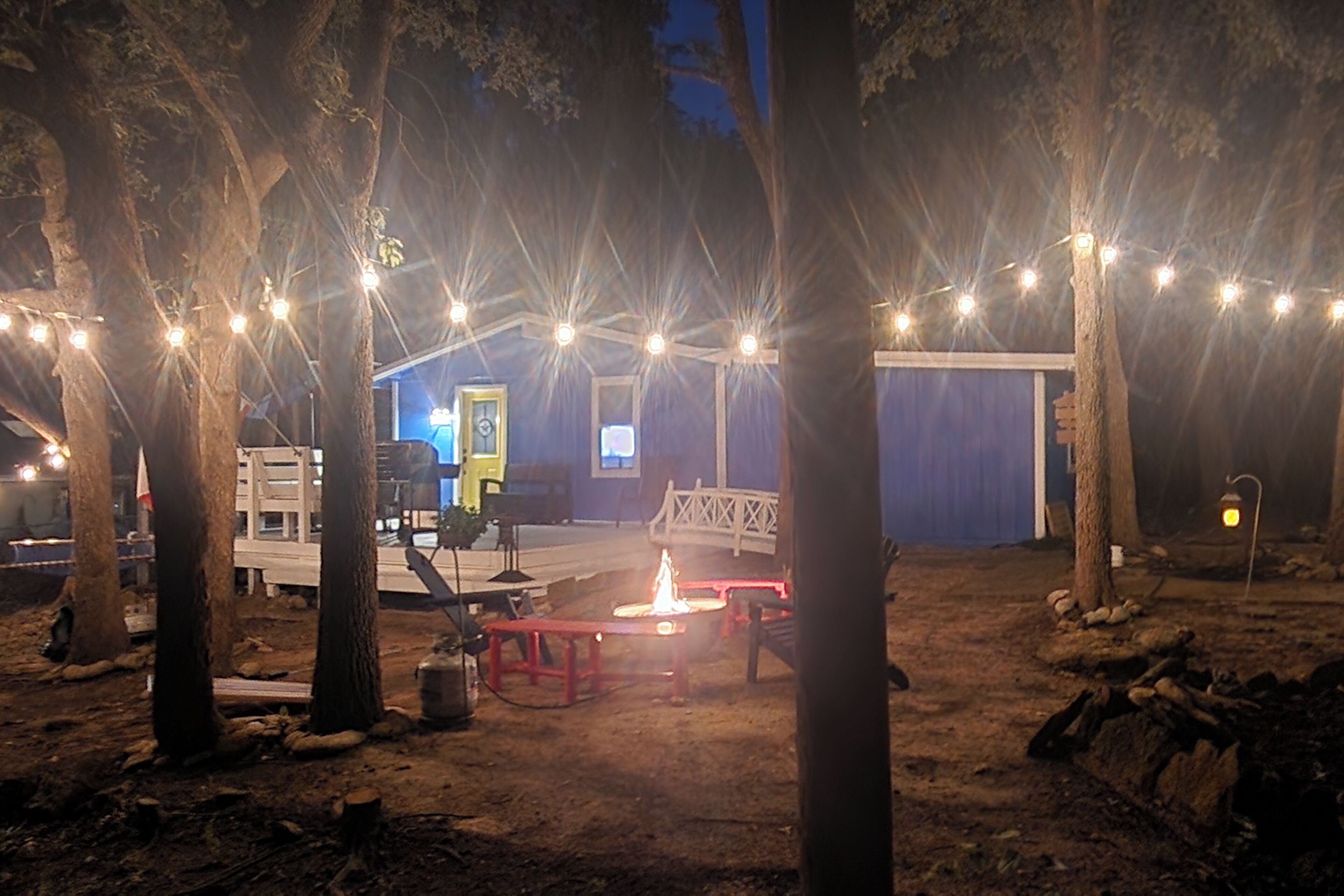 A rustic outdoor area with string lights, a fire pit, and a blue building with a door and windows.