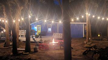 A rustic outdoor area with string lights, a fire pit, and a blue building with a door and windows.