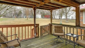 A covered outdoor deck with wooden flooring, a metal railing, and two cushioned chairs and a small table.