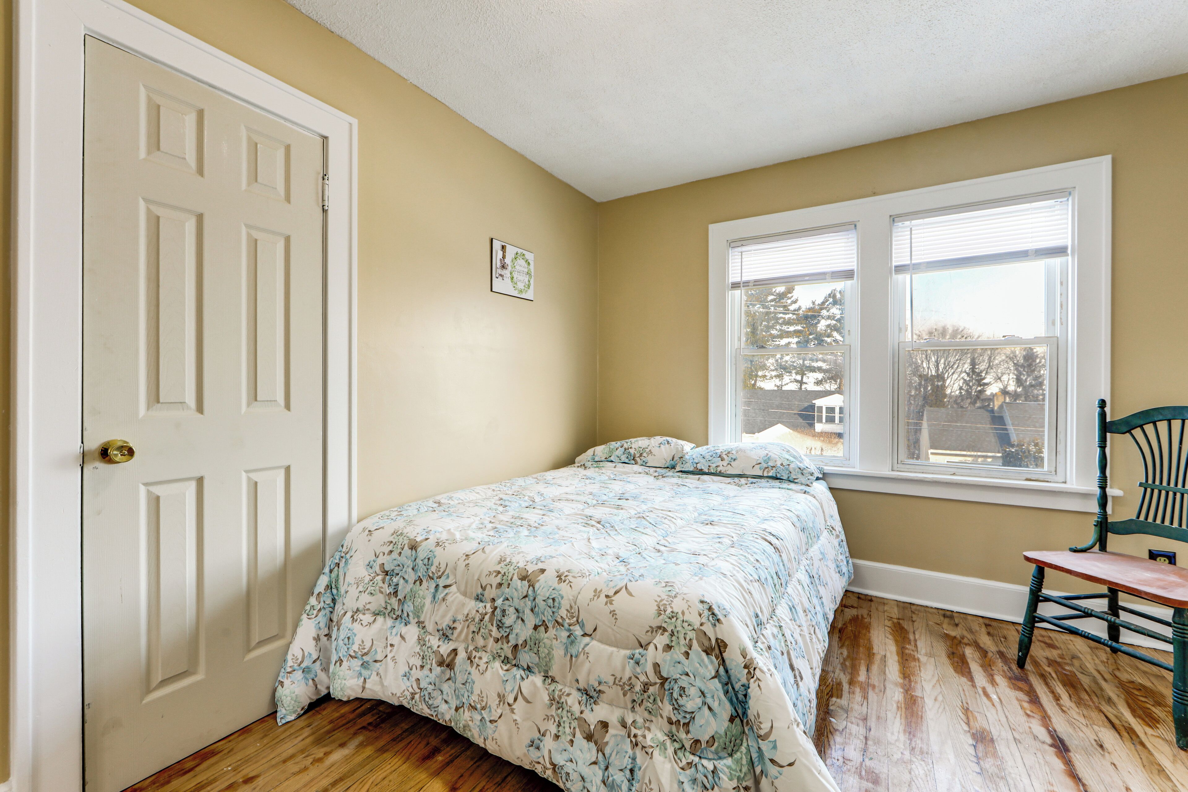 Une chambre à coucher avec une couvre-lit à motifs floraux, un plancher en bois, une porte blanche et une fenêtre avec des stores.