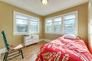 Une chambre avec un plancher en bois, un lit recouvert d’une courtepointe à motifs floraux rouges, une commode blanche et une chaise verte.