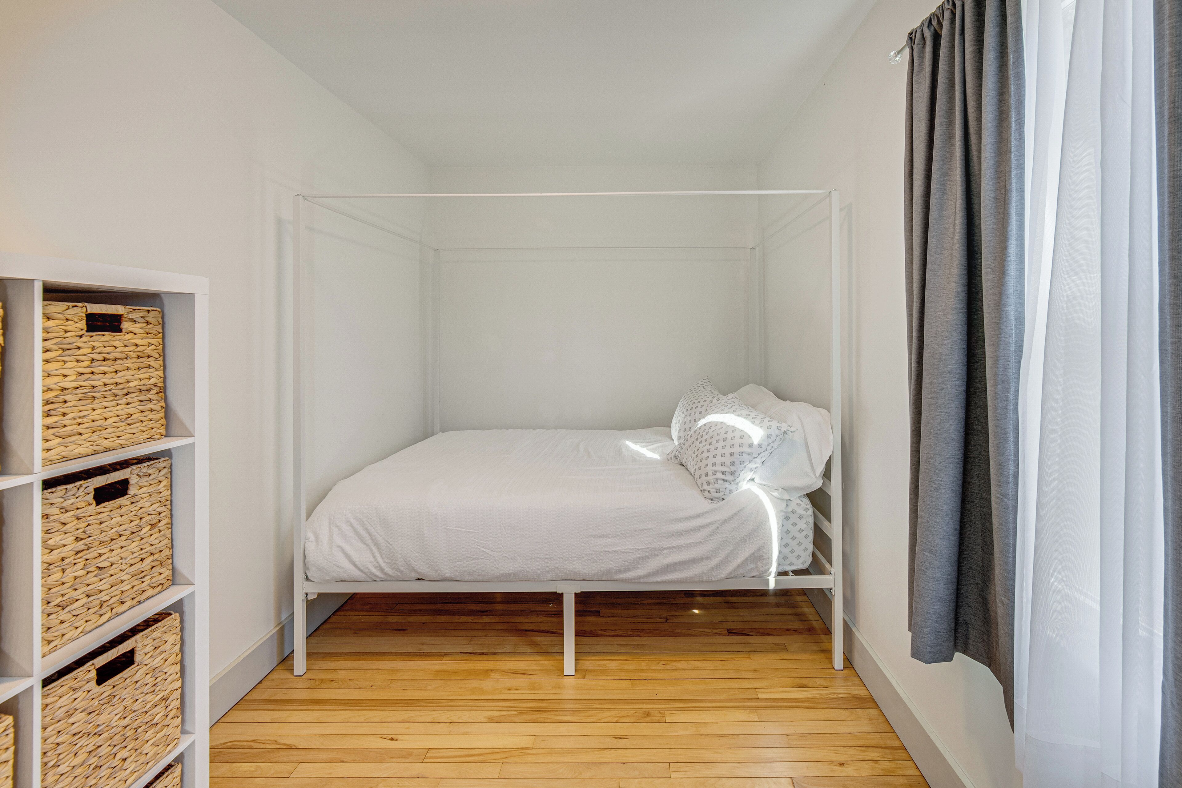 Una habitación pequeña con una cama individual, paredes blancas, suelo de madera, un armario blanco con cestas de mimbre y una ventana con cortinas grises.