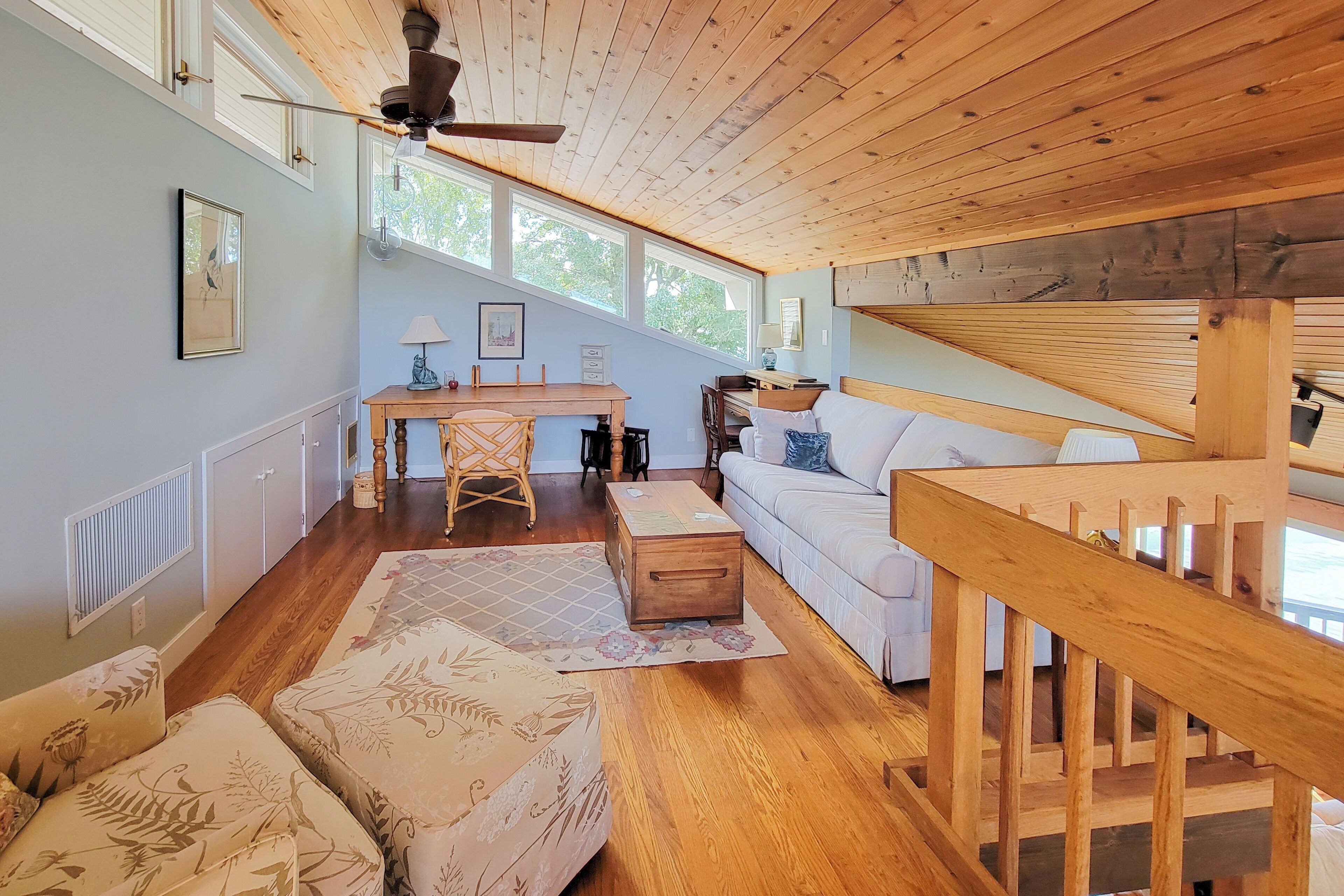 A cozy room with a wooden ceiling, a white sofa, a wooden desk, and a staircase.