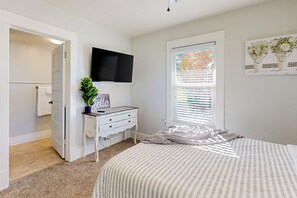 Une chambre à coucher comprenant un lit, une commode, un téléviseur, une fenêtre avec des stores et une porte donnant sur une autre pièce.