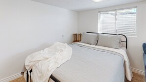 A bedroom with a bed, white walls, a window with blinds, and a ceiling light fixture.