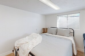 Une chambre à coucher avec un lit, des murs blancs, une fenêtre avec des stores et un luminaire au plafond.