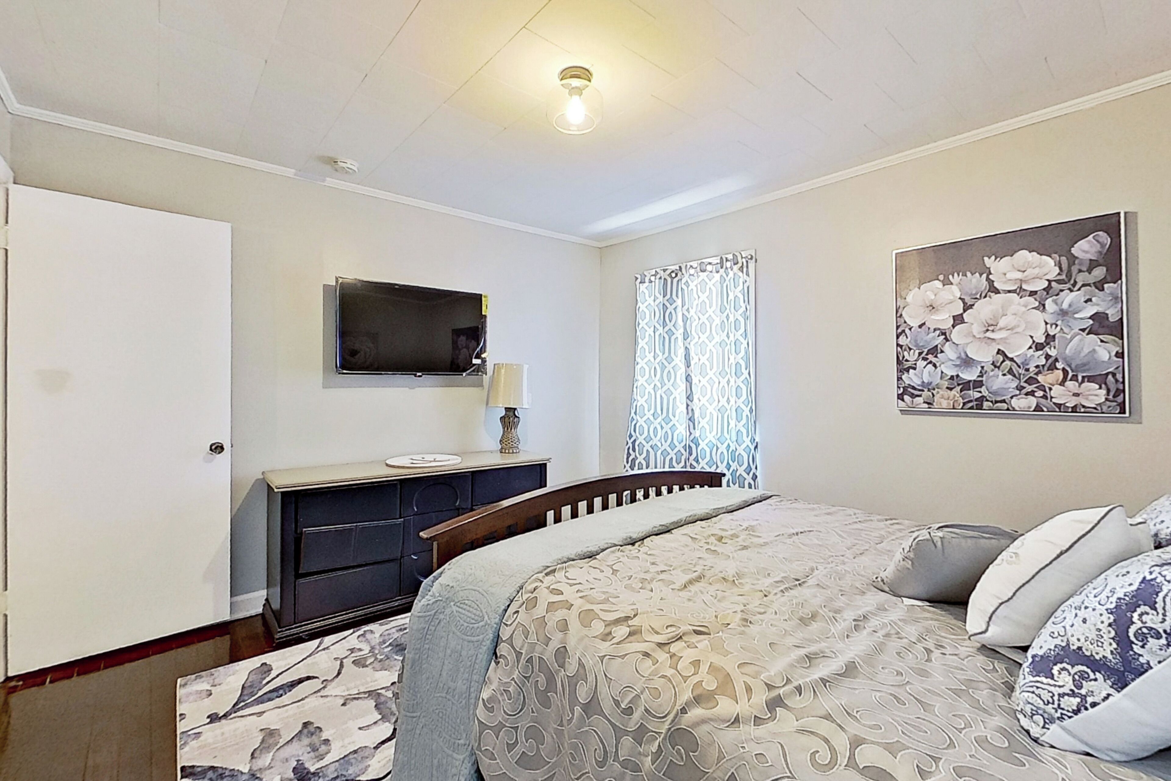 A bedroom with a bed, a dresser, a television, a window with curtains, and a floral painting on the wall.